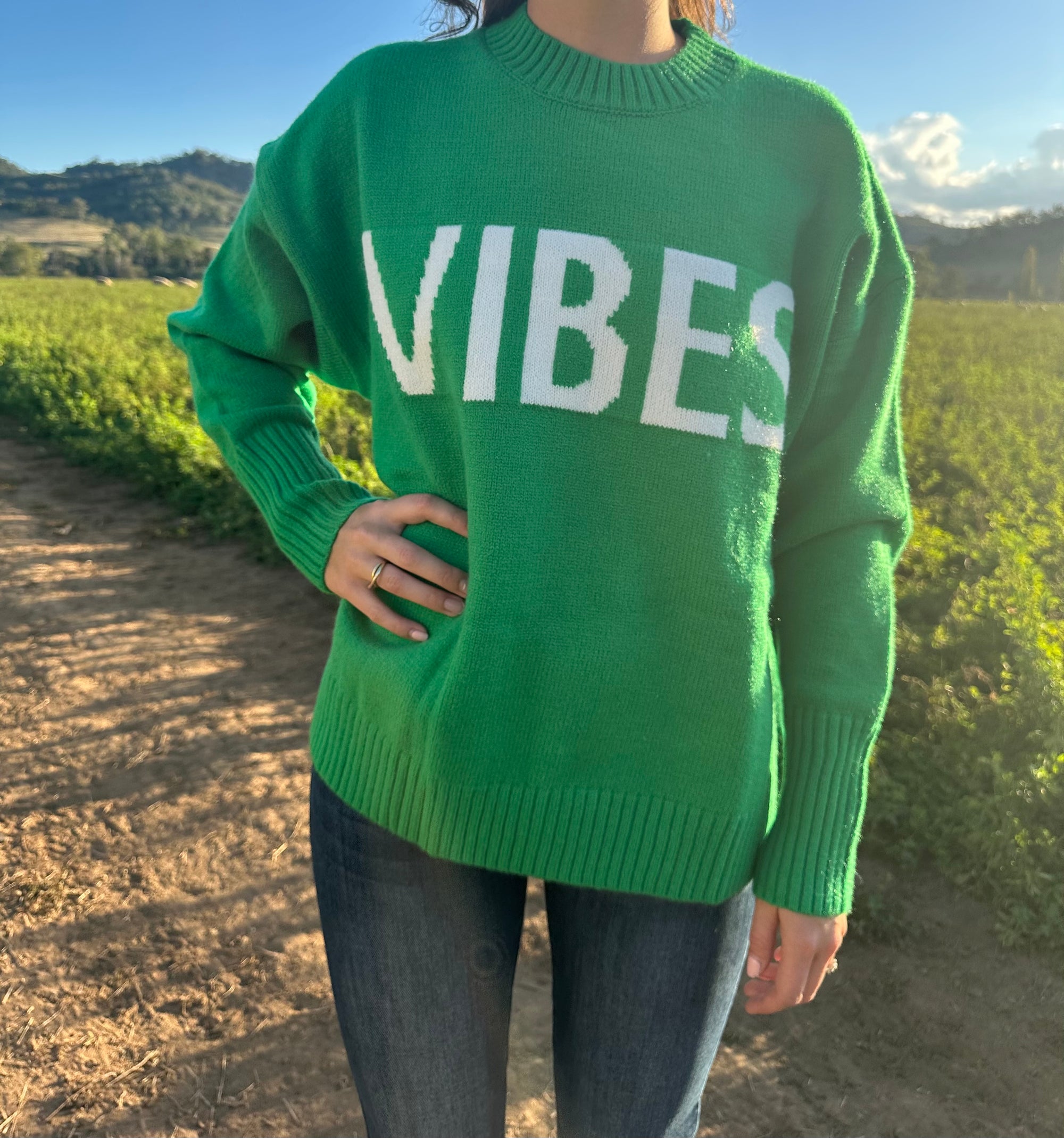 A person wearing a green knit jumper with the word 'VIBES' written across the front in white letters.