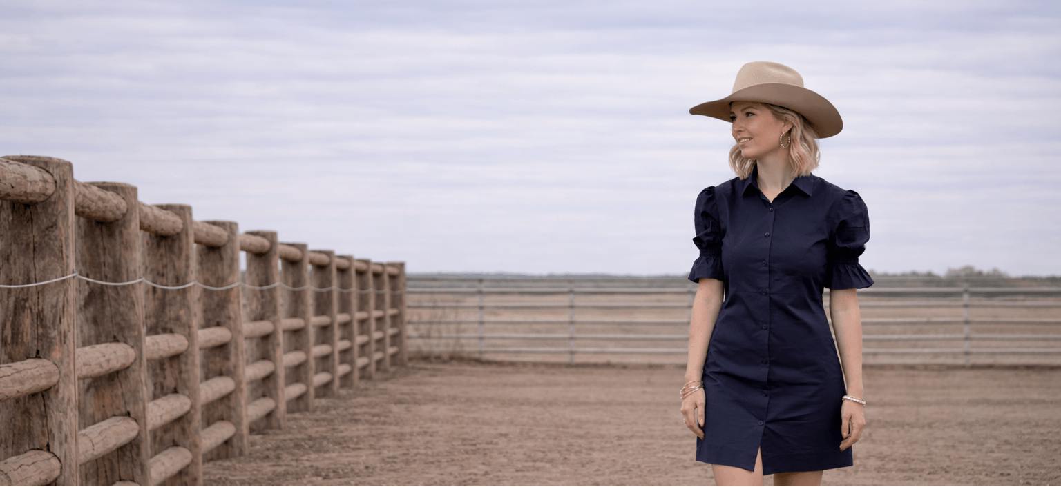 Woman in a navy dress and hat standing in an open field with a wooden fence and cloudy sky.